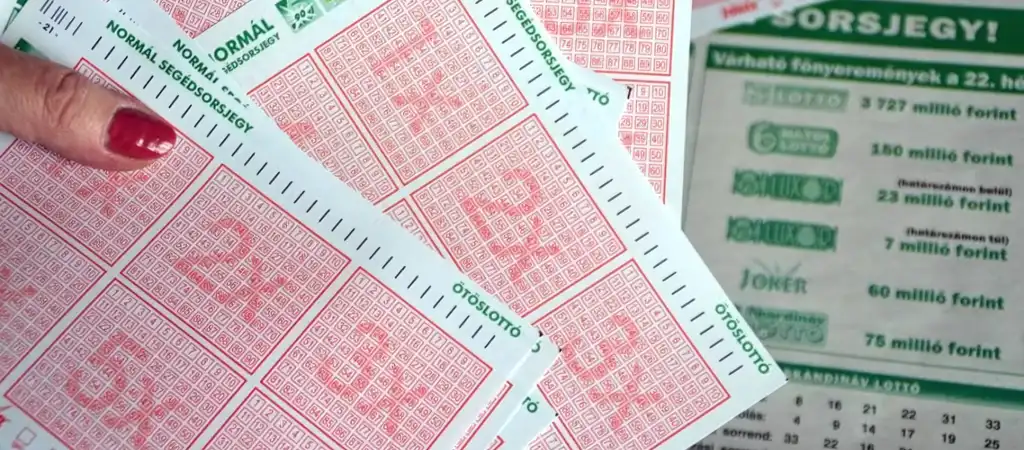 Kéz tart több ötöslottó szelvényt, háttérben a várható főnyeremények listájával.